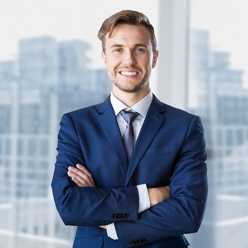 A man in a blue suit stands with arms crossed, smiling confidently against the blurred backdrop of an office building. His light brown hair is neatly styled, and he wears a white shirt with a tie. The scene embodies the spirit of "Empowering Mind's Enriching Lives," blending professionalism with optimism.