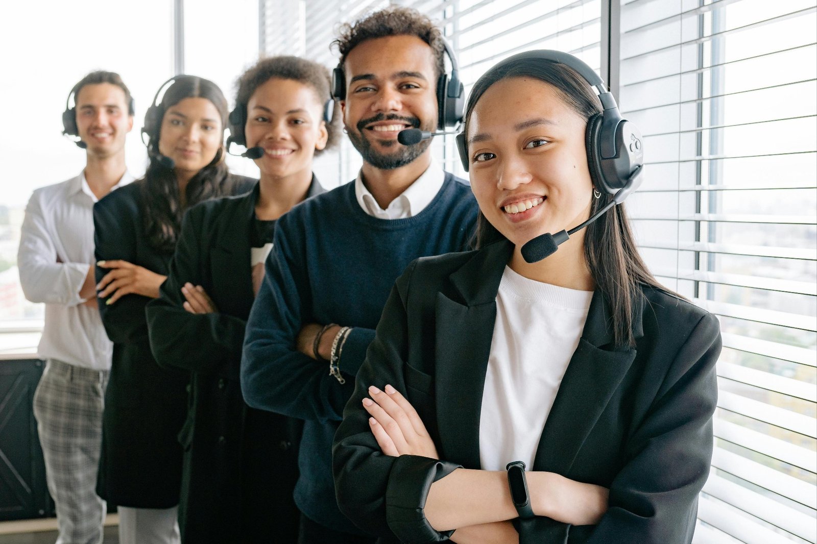 A diverse group of five people stand in an office, wearing headsets and business attire. They are smiling and positioned in a line near large windows, suggesting a modern workplace or customer service environment.